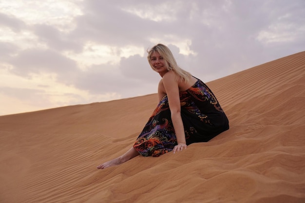 Bella donna bionda in un abito nero è seduta sulla sabbia calda nel deserto