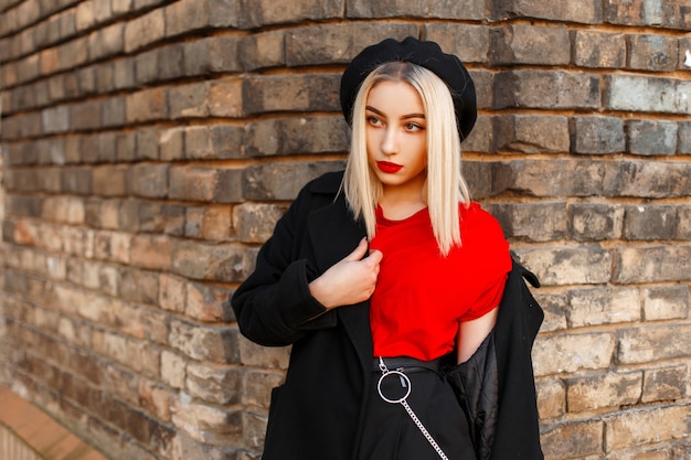 Bella donna bionda in un cappotto di autunno nero vicino a un muro di mattoni d'epoca