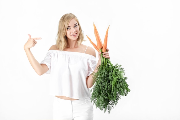 Bella donna bionda in una camicetta bianca mostra una carota fresca con foglie verdi su sfondo bianco. Salute e dieta