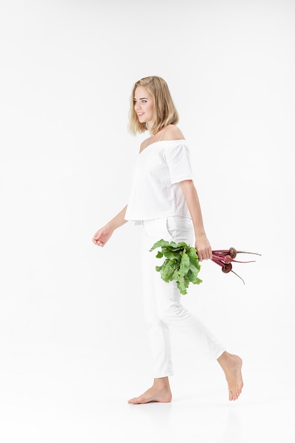 Bella donna bionda in una camicetta bianca tiene una barbabietola con foglie verdi su sfondo bianco. Salute e vitamine