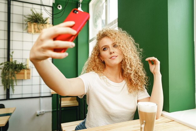 Bella donna bionda riccia che prende selfie mentre è seduta al tavolo in caffetteria