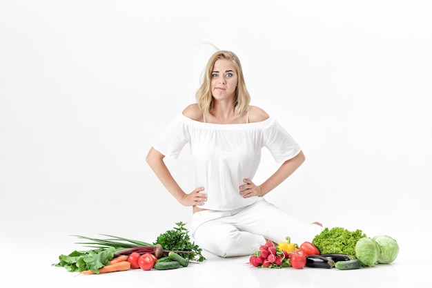 Bella donna bionda sconvolta in abiti bianchi e un sacco di verdure fresche su uno sfondo bianco. dieta