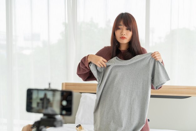 Bella donna blogger asiatica che mostra i vestiti sulla fotocamera per registrare vlog dal vivo nel suo negozio