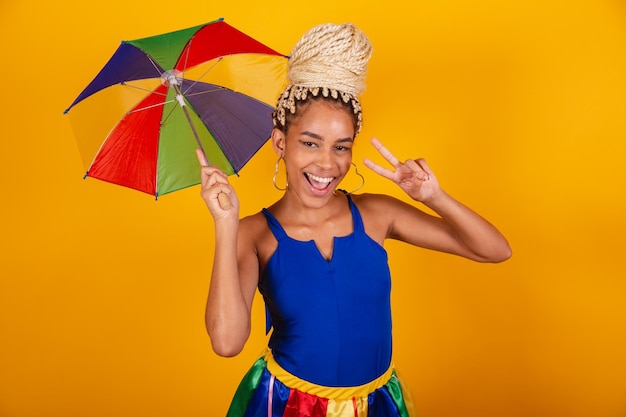 Bella donna brasiliana nera vestita con abiti di carnevale sfondo blu e giallo panino e trecce ombrello frevo Segno di pace e amore
