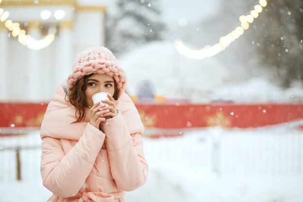 Bella donna bruna che indossa un berretto lavorato a maglia e un cappotto caldo che beve caffè caldo alla fiera invernale. Spazio per il testo