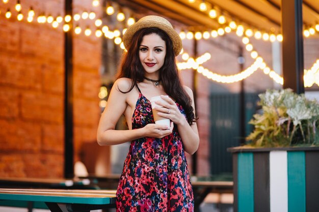 Bella donna bruna in abito estivo lungo e cappello che tiene in mano una tazza di caffè di carta che guarda con sicurezza nella macchina fotografica mentre posa in un accogliente ristorante o in una terrazza all'aperto Concetto di relax