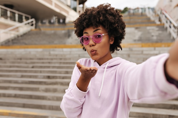 Bella donna bruna piuttosto riccia in elegante felpa con cappuccio rosa e occhiali da sole colorati si bacia e si fa selfie vicino alle scale fuori