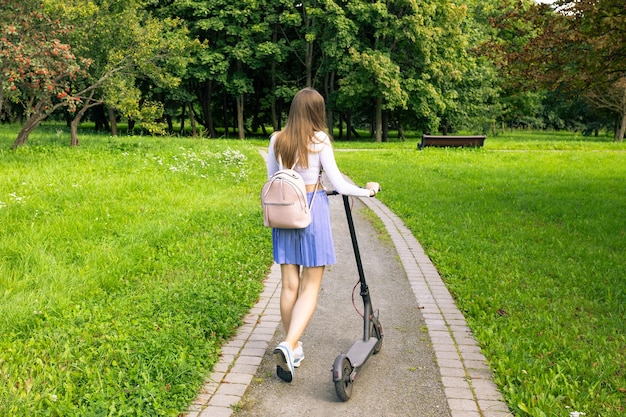 Bella donna cammina lungo il sentiero nel parco e rotola il suo scooter elettrico