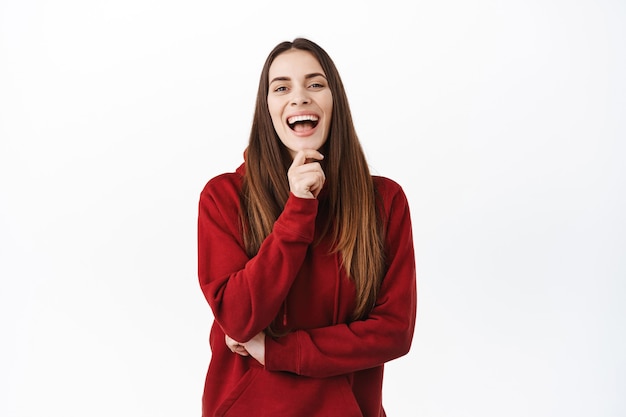 Bella donna candida con lunghi capelli naturali e trucco nudo, ridendo di qualcosa di divertente, ridacchiando e guardando davanti, guardando un evento divertente, in piedi spensierato contro il muro bianco