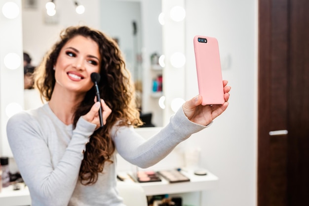 Bella donna castana che prende la foto del selfie mentre applica il trucco davanti allo specchio.