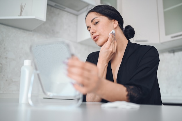 Bella donna castana che rimuove trucco dal suo fronte con lo specchio. Fronte di pulizia della bella ragazza naturale carina bellezza con spugna di cotone. Cosmetologia e spa, problemi di cura della pelle, concetto di trattamento dell'acne