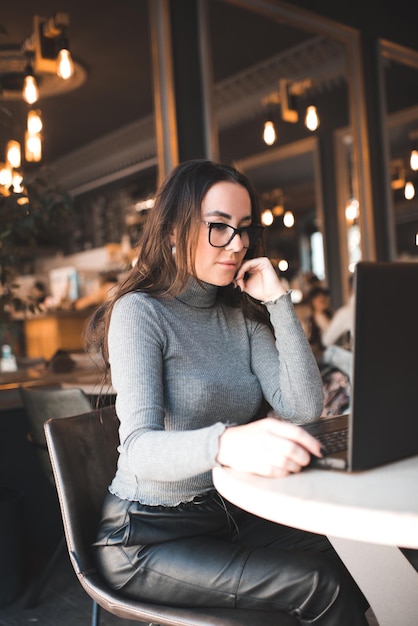 Bella donna castana con i capelli ricci che si siede nella caffetteria che lavora al computer portatile sopra le luci d'ardore