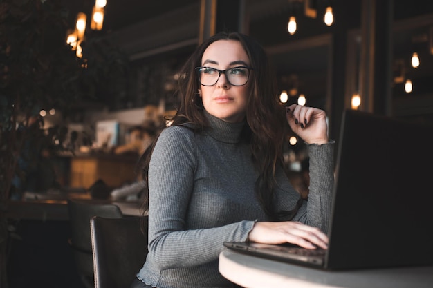 Bella donna castana con i capelli ricci che si siede nella caffetteria che lavora al computer portatile sopra le luci d'ardore