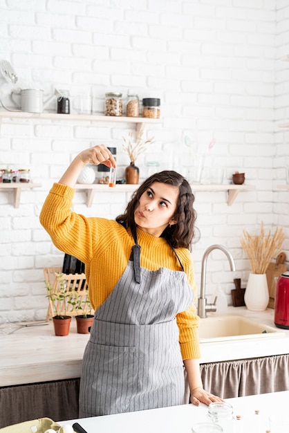 Bella donna castana in maglione giallo e grembiule grigio colorare le uova di Pasqua in cucina che tiene la provetta con colore arancione