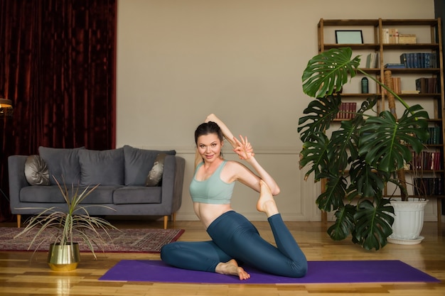 Bella donna castana in un top e leggings esegue pose di yoga sulla stuoia in una stanza a casa