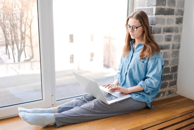 Bella donna casuale felice in vetri che lavora al computer portatile che si siede sul davanzale