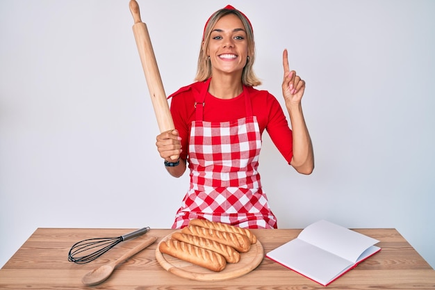 Bella donna caucasica che indossa il grembiule da panettiere che fa il pane fatto in casa sorridendo con un'idea o una domanda che punta il dito con la faccia felice numero uno