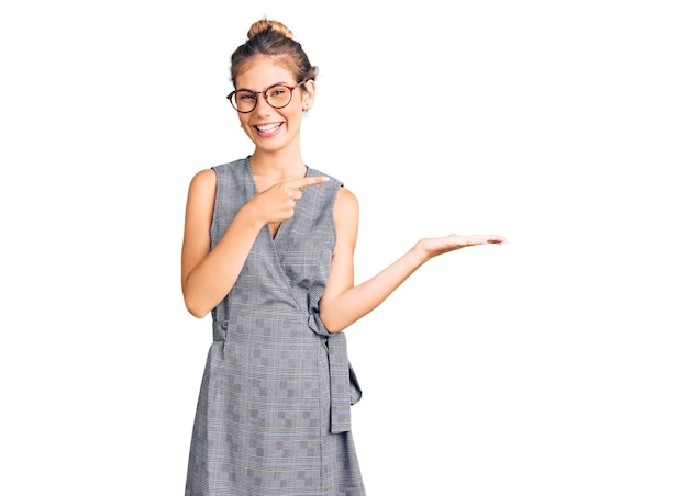 Bella donna caucasica con i capelli biondi che indossa abiti da lavoro e occhiali stupiti e sorridendo alla telecamera mentre si presenta con la mano e indica con il dito