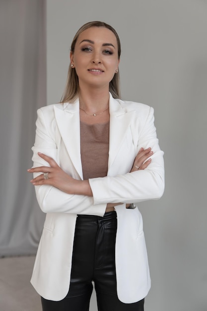 Bella donna caucasica con una giacca bianca e pantaloni neri che sorride alla telecamera su uno sfondo grigio in uno studio fotografico