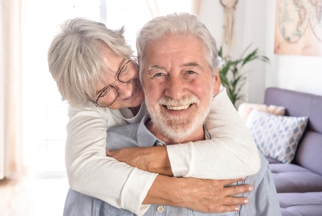 Bella donna caucasica senior che abbraccia amorevolmente il marito seduto Sorridente coppia di anziani dai capelli bianchi guardando la fotocamera