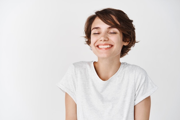 Bella donna caucasica sognante sorridente con gli occhi chiusi, in piedi rilassata e felice contro il muro bianco white