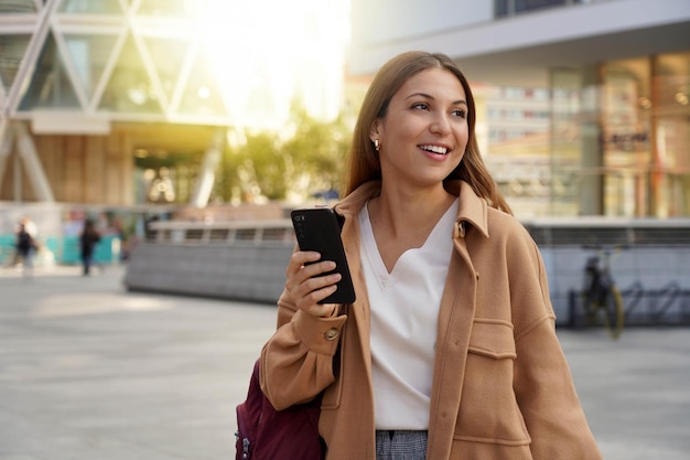 Bella donna che cammina nella città moderna utilizzando il telefono cellulare guardando il lato all'ora del tramonto