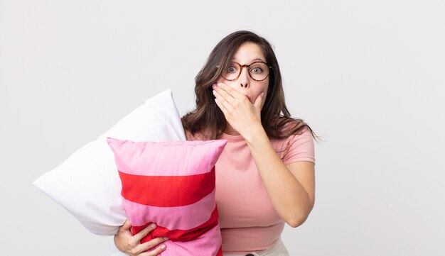 Bella donna che copre la bocca con le mani con un pigiama scioccato e tiene un cuscino