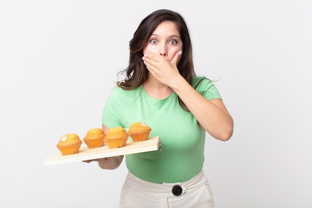 Bella donna che copre la bocca con le mani con un vassoio scioccato e con in mano un muffin