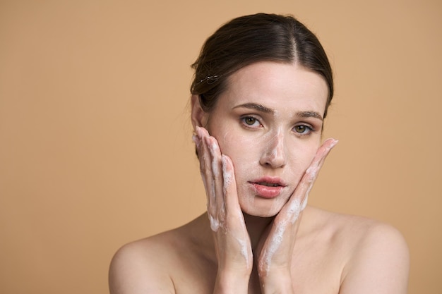 Bella donna che guarda l'obbiettivo mentre applica la schiuma detergente sul viso isolata sul beige