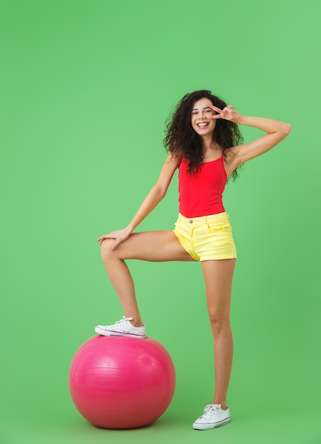 bella donna che indossa abiti estivi facendo esercizi con palla fitness durante l'aerobica contro il muro verde