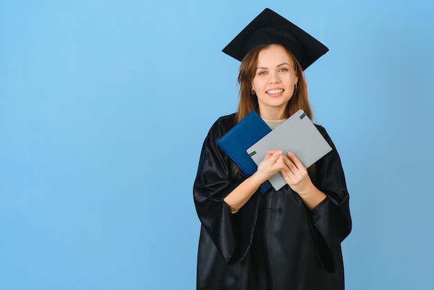 Bella donna che indossa il berretto di laurea e l'abito da cerimonia in possesso di laurea che sembra positivo e felice in piedi e sorride con un sorriso fiducioso.