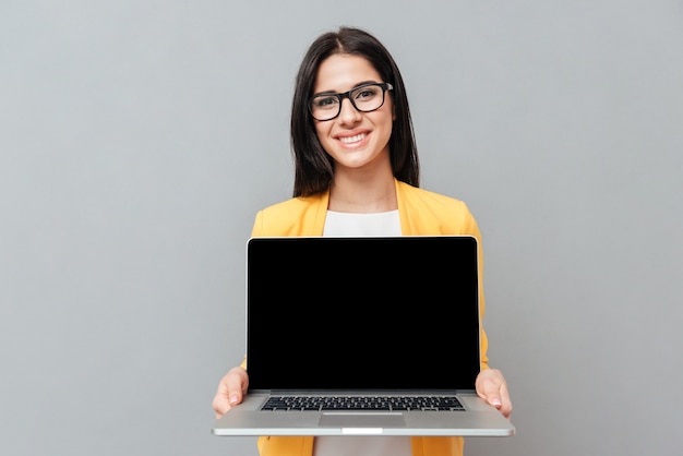 Bella donna che indossa occhiali e vestita con una giacca gialla che mostra il display del laptop in primo piano su una superficie grigia.