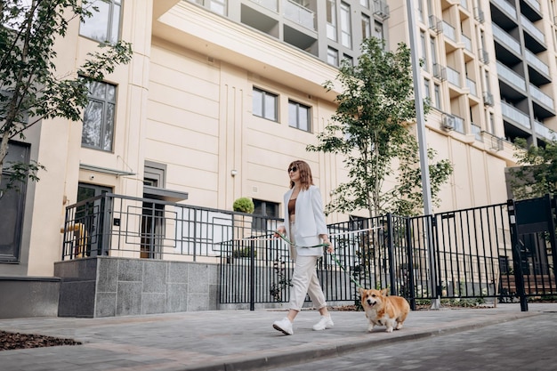Bella donna che indossa un abito bianco e occhiali da sole che cammina per strada con il cane Welsh Corgi Pembroke