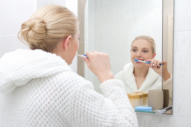 Bella donna che lava i denti e si guarda allo specchio in bagno