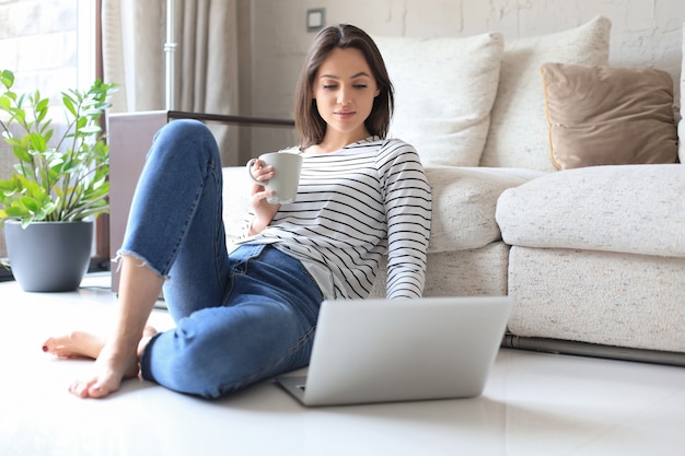 Bella donna che lavora con il computer portatile mentre è seduta sul pavimento a casa e beve caffè.