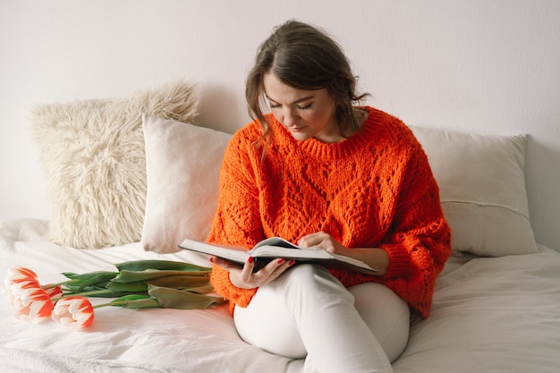 Bella donna che legge il libro a letto a casa. La donna si rallegra dei tulipani.