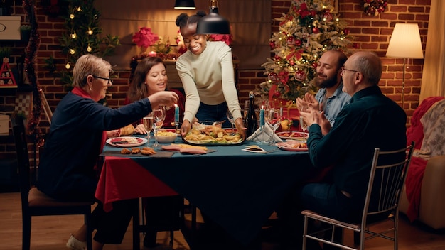 Bella donna che porta il piatto principale al tavolo da pranzo di Natale. I membri della famiglia allegri e festivi si sono riuniti a casa per celebrare le vacanze invernali con cibo tradizionale cucinato in casa.