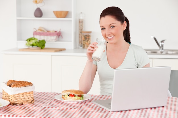 Bella donna che posa con un bicchiere di latte mentre si rilassa con il suo computer portatile a ora di pranzo