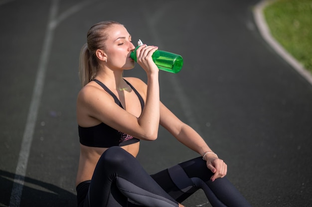 Bella donna che riposa dopo l'allenamento allo stadio, la ragazza si sedette per rilassarsi, bevendo acqua dalla bottiglia verde.