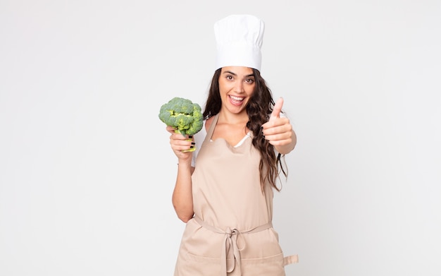 Bella donna che si sente orgogliosa, sorride positivamente con il pollice in alto indossando un grembiule e tenendo in mano un broccolo