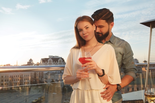 Bella donna che sorride alla telecamera mentre il suo ragazzo la abbraccia sul balcone sul tramonto