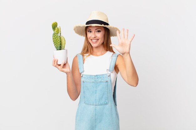 Bella donna che sorride e sembra amichevole, mostra il numero cinque e tiene in mano una pianta decorativa di cactus