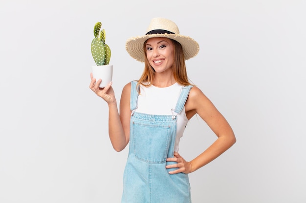 Bella donna che sorride felicemente con una mano sull'anca e sicura di sé e tiene in mano una pianta decorativa di cactus