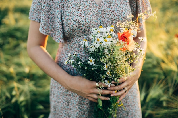 Bella donna che tiene un bouquet di fiori selvatici vicino alla luce del tramonto in un campo d'orzo Femmina elegante che si rilassa la sera in campagna estiva e raccoglie fiori Momento tranquillo atmosferico