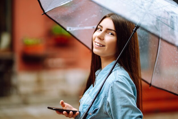 Bella donna che utilizza smartphone su strada in caso di pioggia sorridente tenendo ombrello Pioggia estiva
