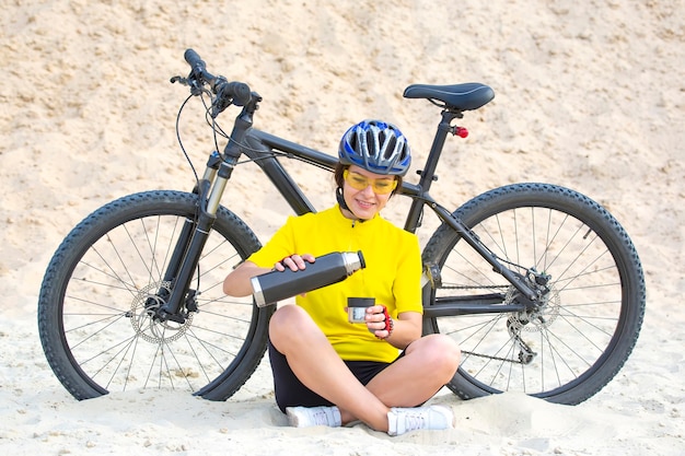 Bella donna ciclista in giallo con tè e thermos in mano sullo spazio di sabbia. Sport e ricreazione.