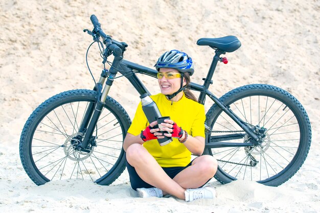 Bella donna ciclista versa il tè da un thermos sullo sfondo di una bicicletta nella sabbia. Sport e ricreazione. Hobby e salute.