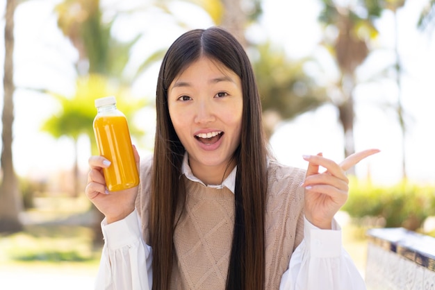 Bella donna cinese che tiene un succo d'arancia all'aperto sorpreso e punta il dito di lato