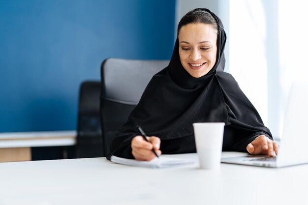 Bella donna con abito abaya che lavora al suo computer. Impiegata di mezza età al lavoro in un ufficio commerciale a Dubai. Concetto di culture e stile di vita mediorientali