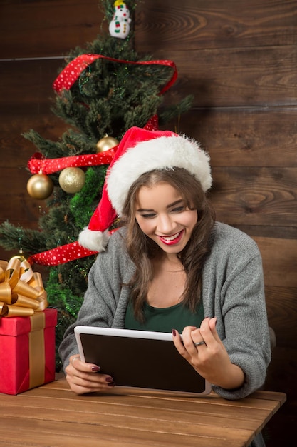 Bella donna con cappello da Babbo Natale che utilizza un tablet PC mentre trascorre il fine settimana a casa vicino all'albero di Capodanno.